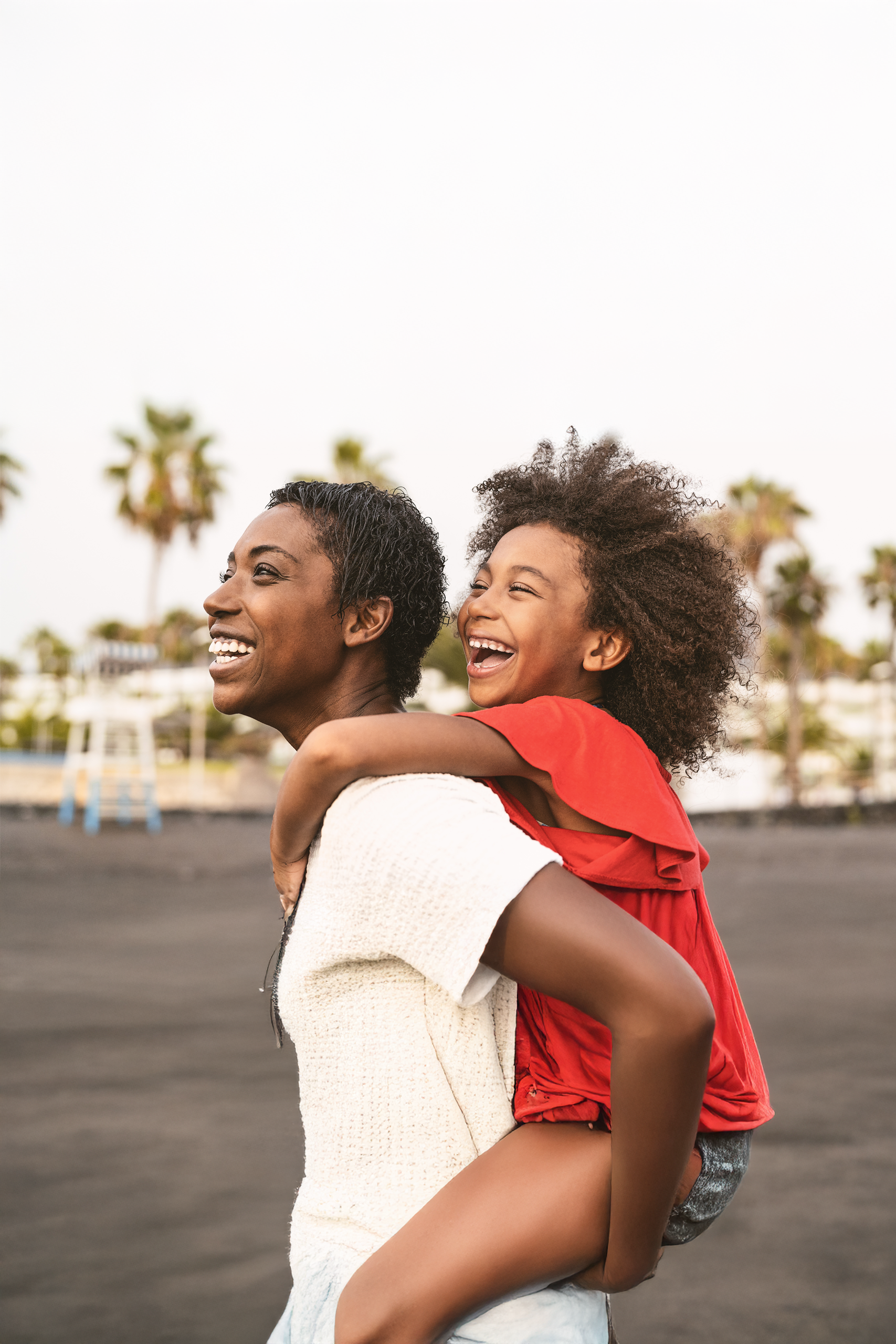 FLTDSO image At an outdoor basketball court with apartment buildings in the background, a parent and child share a joyful moment as the adult lifts the child up to reach toward the basketball. The child, wearing a white t-shirt and grey shorts, has curly natural hair and reaches up enthusiastically while holding a basketball. The adult, wearing a beige t-shirt, smiles warmly while supporting the child